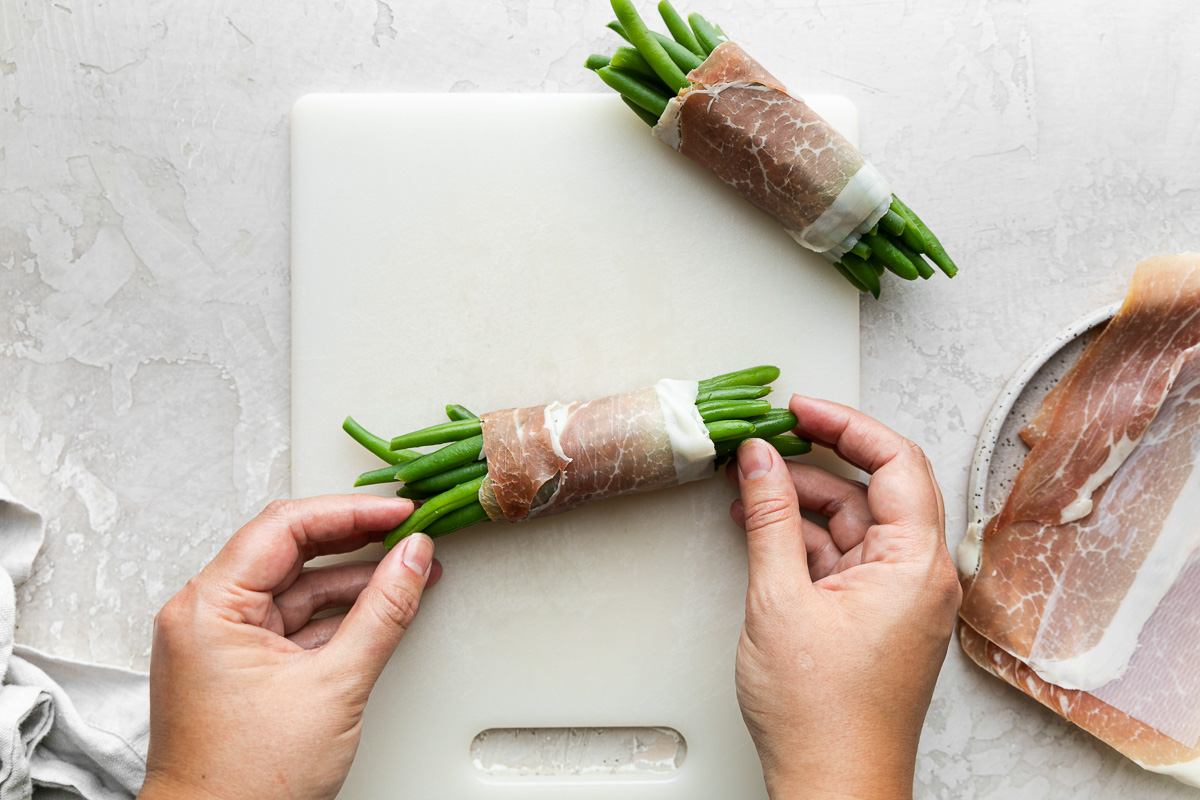 A woman's hands hold a finished prosciutto-wrapped green bean bundle atop a while cutting board, while another bundle rests above it also atop the cutting board. To the right of the cutting board is a gray speckled ceramic plate holding sheets of prosciutto and to the left of the cutting board rests a light gray linen napkin. The cutting board, napkin, and plate all sit atop a creamy white plaster surface.