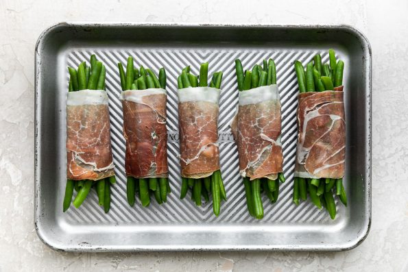 Five finished prosciutto-wrapped green bean bundles arranged on a small aluminum baking sheet. The baking sheet sits atop a creamy white plaster surface.