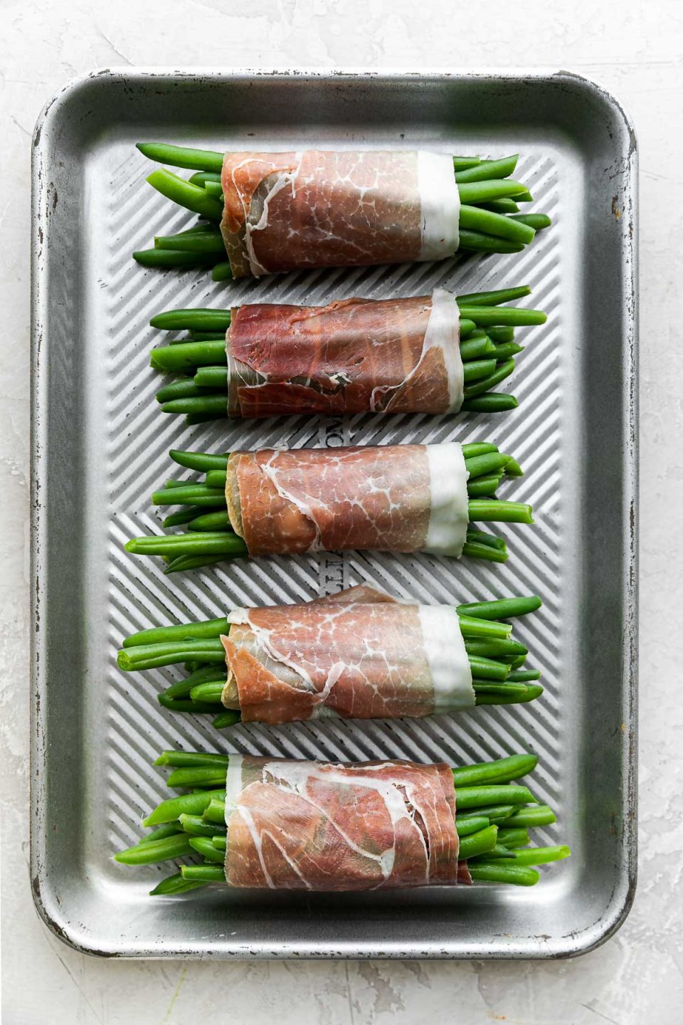 Five finished prosciutto-wrapped green bean bundles arranged on a small aluminum baking sheet. The baking sheet sits atop a creamy white plaster surface.