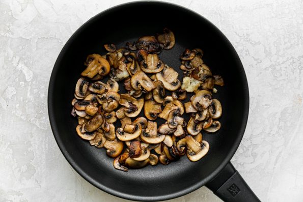 A black skillet is filled with golden brown cooked mushrooms. The skillet sits atop a creamy white plaster surface.