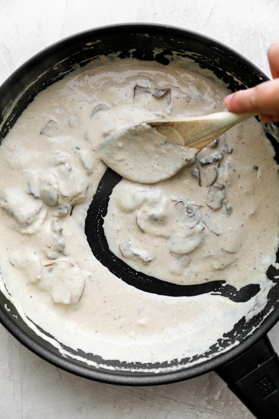 A black skillet is filled with a creamy mushroom sauce and a woman's hand holds a wooden spoon as she works to stir the sauce. The skillet sits atop a creamy white plaster surface.