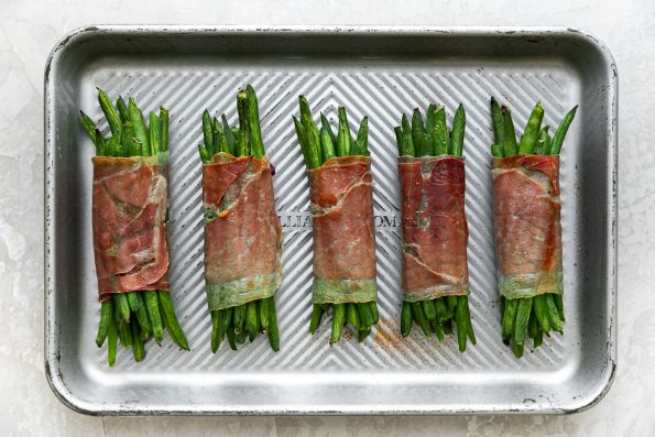 Five roasted prosciutto-wrapped green bean bundles arranged on a small aluminum baking sheet. The baking sheet sits atop a creamy white plaster surface.