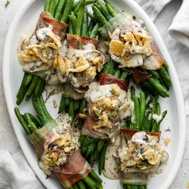 Prosciutto-wrapped green bean bundles arranged on a white serving platter and topped with creamy mushroom sauce and french fried onions. The serving platter sits atop a creamy white plaster surface and light gray linen napkin is tucked underneath the platter.