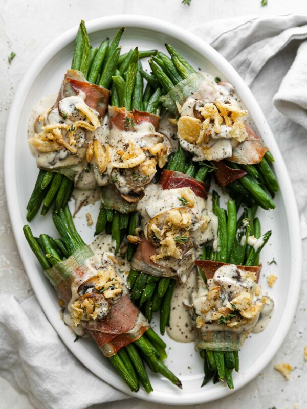 Prosciutto-wrapped green bean bundles arranged on a white serving platter and topped with creamy mushroom sauce and french fried onions. The serving platter sits atop a creamy white plaster surface and light gray linen napkin is tucked underneath the platter.