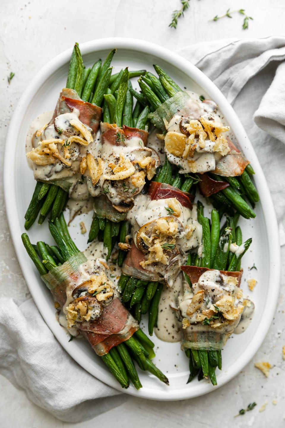Prosciutto-wrapped green bean bundles arranged on a white serving platter and topped with creamy mushroom sauce and french fried onions. The serving platter sits atop a creamy white plaster surface and light gray linen napkin is tucked underneath the platter.