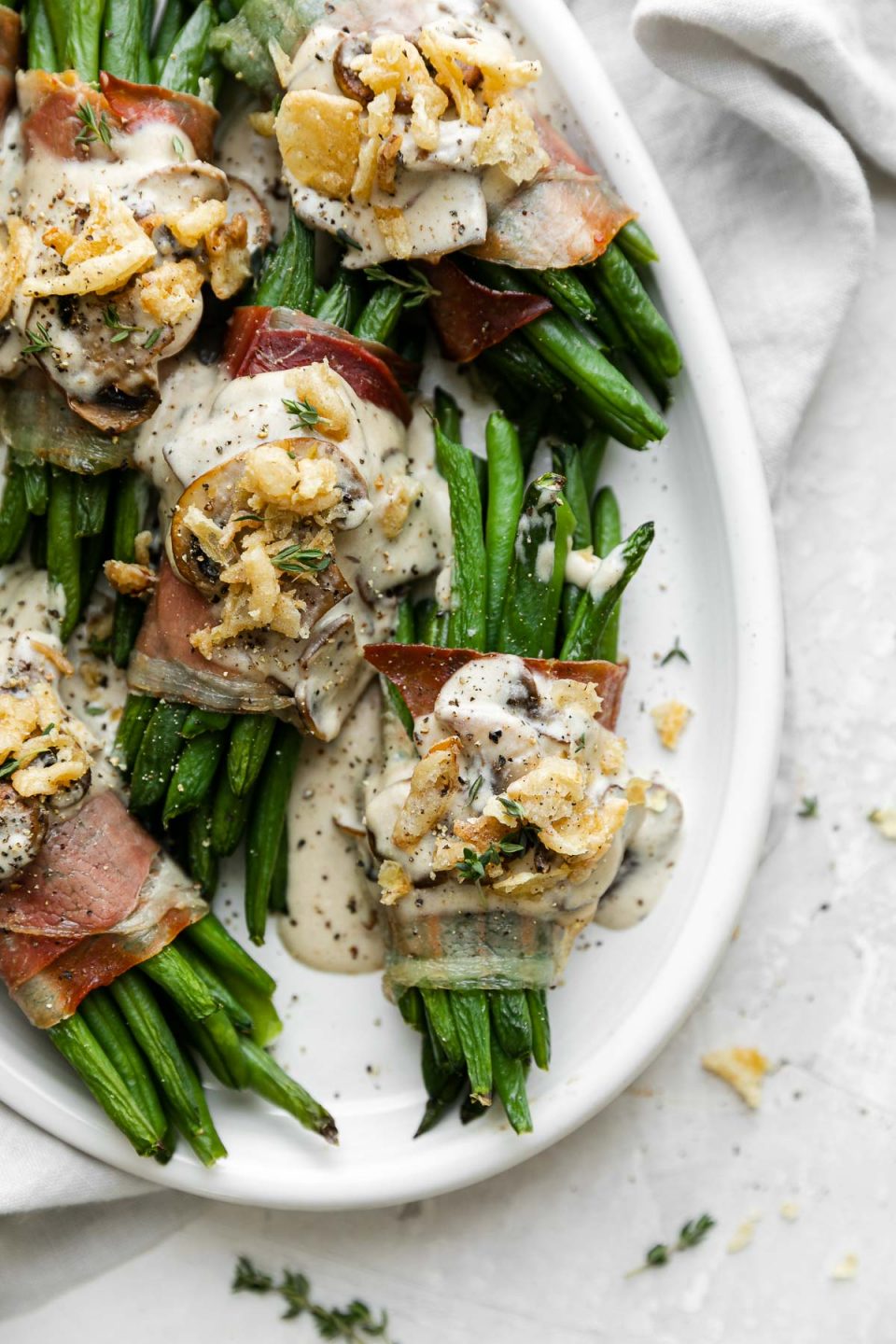 A close up of prosciutto-wrapped green bean bundles arranged on a white serving platter and topped with creamy mushroom sauce and french fried onions. The serving platter sits atop a creamy white plaster surface and light gray linen napkin is tucked underneath the platter.