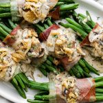 A close up of multiple prosciutto-wrapped green bean bundles arranged on a white serving platter and topped with creamy mushroom sauce and french fried onions. A light gray linen napkin is tucked underneath the serving platter.