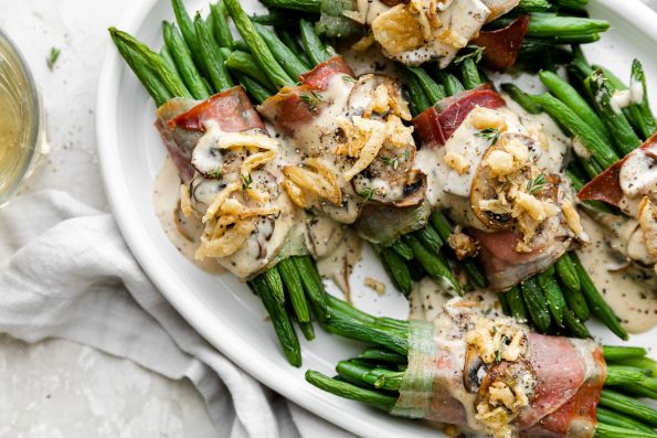 A close up of multiple prosciutto-wrapped green bean bundles arranged on a white serving platter and topped with creamy mushroom sauce and french fried onions. A light gray linen napkin is tucked underneath the serving platter and single glass of white wine sits alongside the platter.