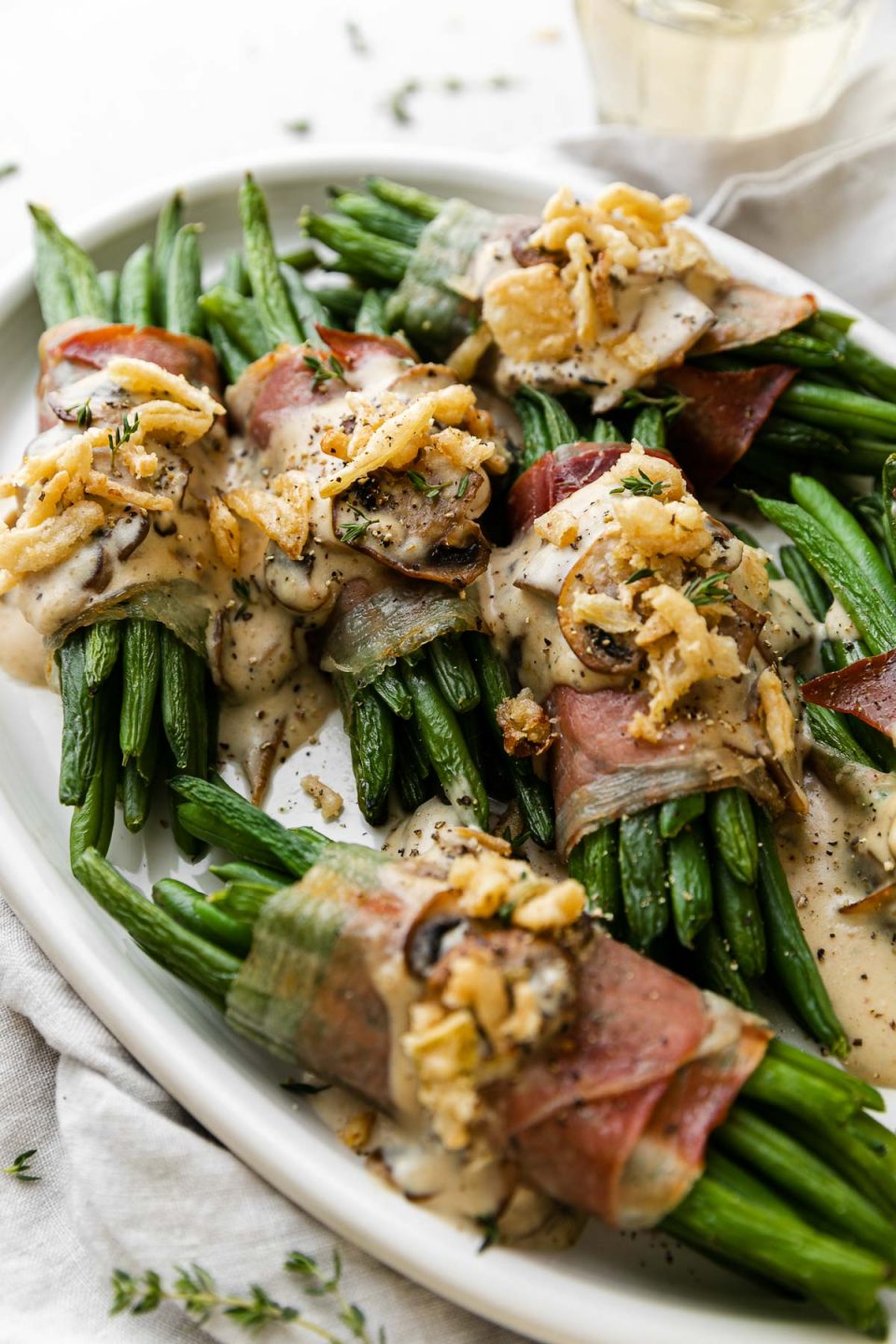 An overhead shot of ​multiple prosciutto-wrapped green bean bundles arranged on a white serving platter and topped with creamy mushroom sauce and french fried onions. A light gray linen napkin is tucked underneath the serving platter and a glass of white wine sits behind the platter in the background.
