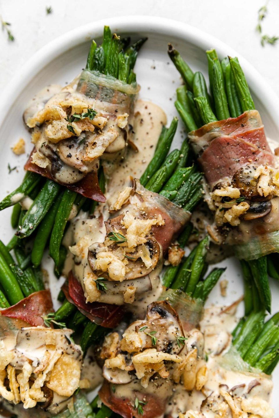 An overhead shot of ​multiple prosciutto-wrapped green bean bundles arranged on a white serving platter and topped with creamy mushroom sauce and french fried onions.
