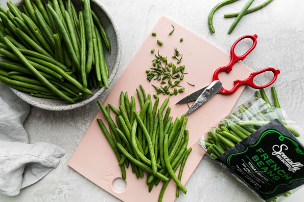 A pile of trimmed green beans rest on a pink cutting board next to a pair of red kitchen shears and a pile of discarded green bean ends that have been snipped off using the shears. A gray speckled ceramic bowl sits to the left of the cutting board filled with trimmed and cleaned green beans along with a light gray linen napkin and a bag of Specially Selected French green beans sits to the right of the cutting board filled with green beans that have yet to be trimmed.