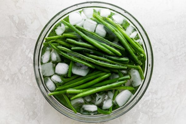 Blanched green beans rest in clear glass mixing bowl filled with ice water. The mixing bowl sits atop a creamy white plaster surface.
