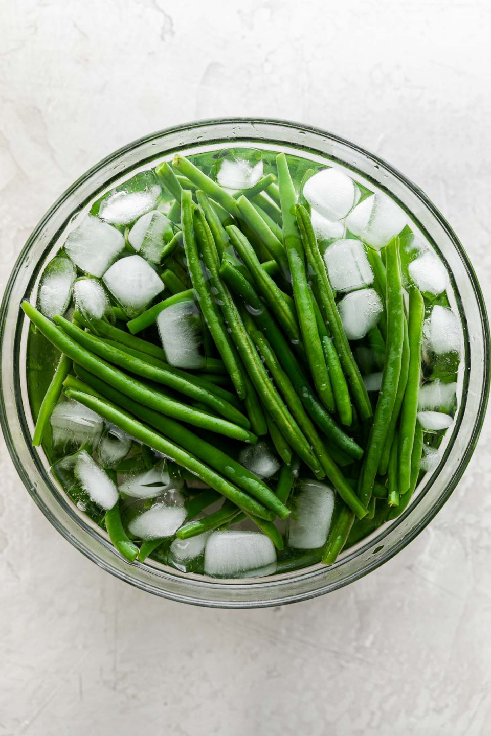 Blanched green beans rest in clear glass mixing bowl filled with ice water. The mixing bowl sits atop a creamy white plaster surface.