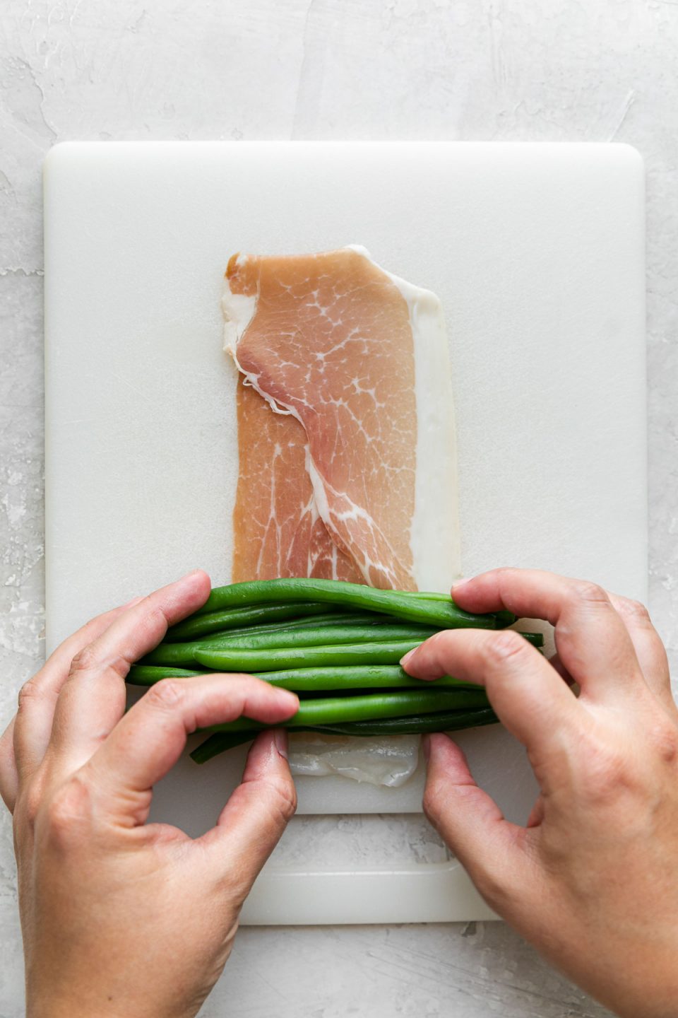 A woman's hands work to gather a pile of neatly arranged green beans running in the same direction and place the pile at the end of a single piece of prosciutto that is resting atop a white cutting board. The cutting board sits atop a creamy white plaster surface.