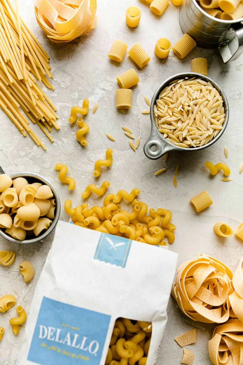 An overhead shot of raw uncooked dried pasta of different varieties: cavatappi, orzo, mezzi rigatoni, pappardelle nests, shellbows, and spaghetti from DeLallo Foods arranged on a creamy white plaster surface. The pasta is scattered about and an opened package of DeLallo dried Cavatappi pasta spills out onto the surface.
