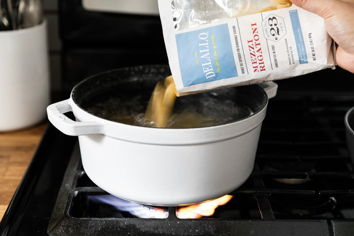 A woman's hand holds a package of raw uncooked DeLallo Mezzi Rigatoni pasta and is pouring it into a white dutch oven atop a gas stovetop.
