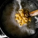 A spider strainer is submerged into a white dutch oven filled with boiling water and half-cooked pasta. The spider strainer is used to pick up a handful of Mezzi Rigatoni pasta noodles to check for doneness. The dutch oven sits atop a gas stovetop range.