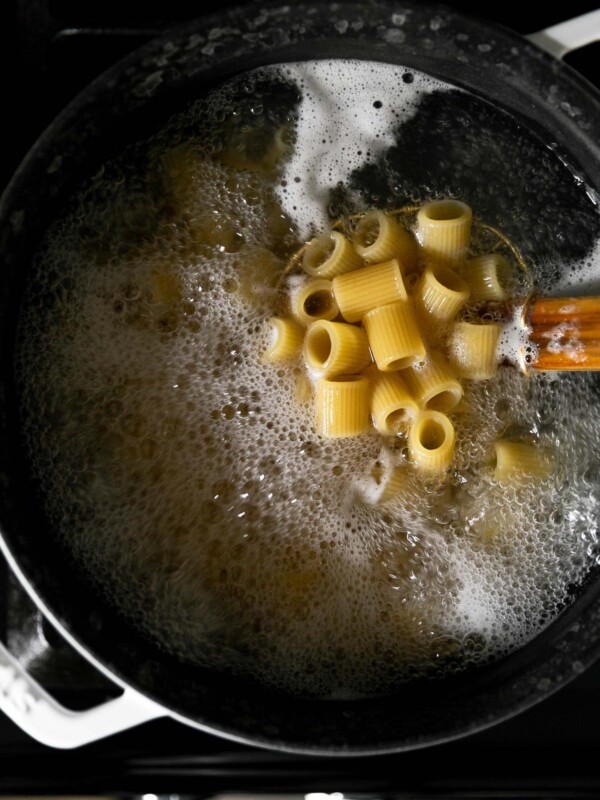 A spider strainer is submerged into a white dutch oven filled with boiling water and half-cooked pasta. The spider strainer is used to pick up a handful of Mezzi Rigatoni pasta noodles to check for doneness. The dutch oven sits atop a gas stovetop range.