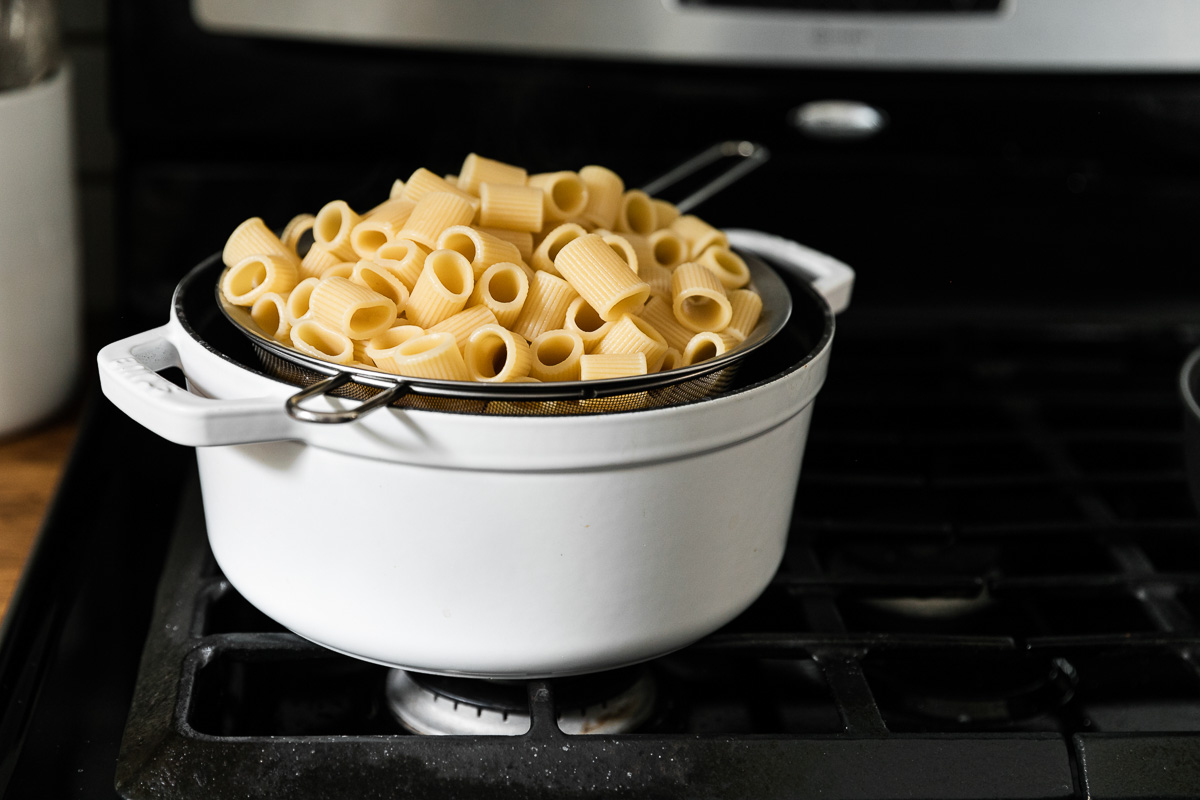 A mesh strainer full of al dente cooked Mezzi Rigatoni sits atop a white dutch oven. The dutch oven rests atop a gas stovetop.