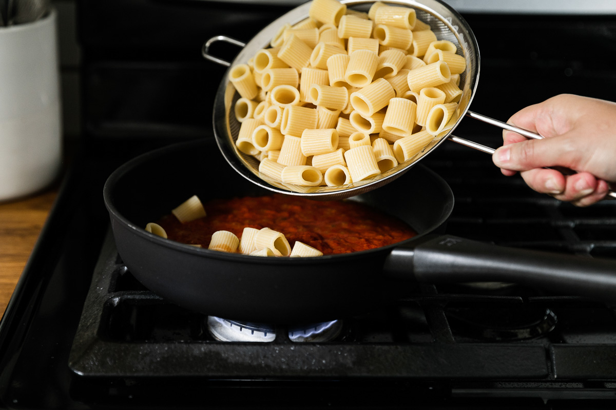 Al dente Mezzi Rigatoni pasta is added to a skillet filled with pasta sauce from a small mesh strainer. A woman's hand holds the strainer full of drained pasta over the skillet and the skillet rests on a gas stovetop.