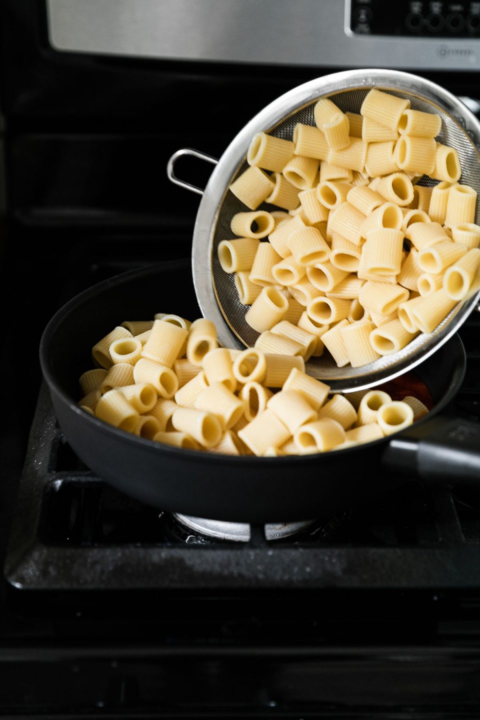 Al dente Mezzi Rigatoni pasta is added to a skillet filled with pasta sauce from a small mesh strainer. The skillet rests on a gas stovetop.