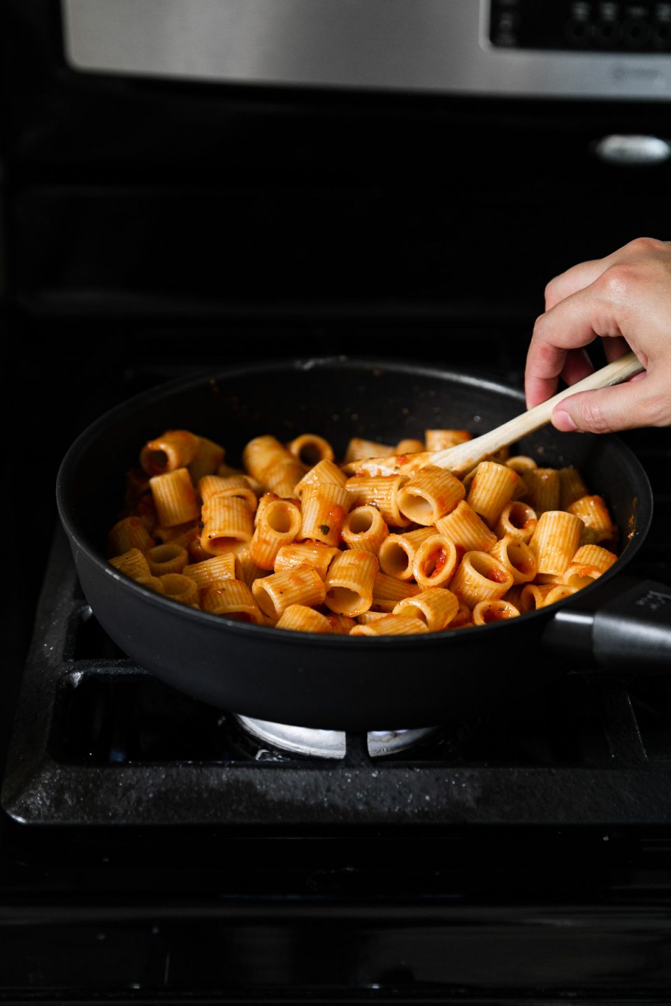 Cooked DeLallo Mezzi Rigatoni in a skillet tossed and combined with pasta sauce a woman's hand holds the skillet and a wooden spoon in her other hand to stir the pasta. The skillet rests atop a gas stovetop range.