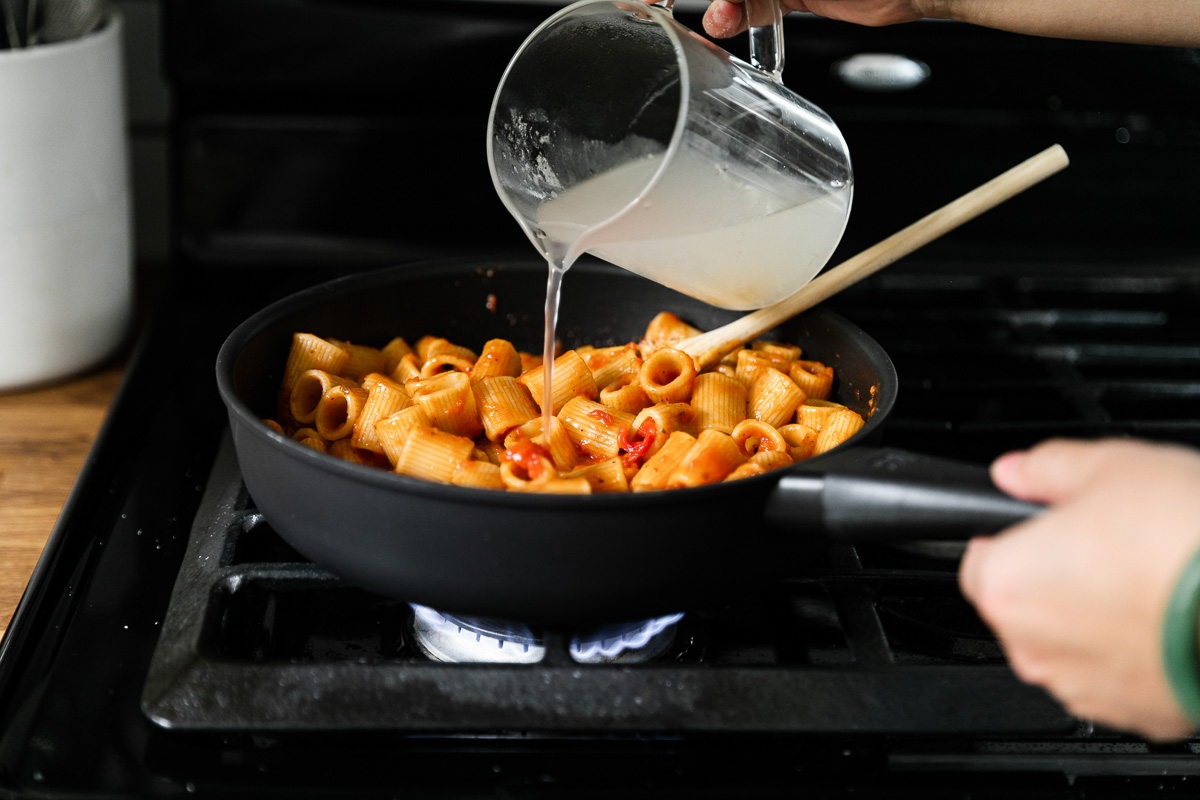 Cooked DeLallo Mezzi Rigatoni in a skillet tossed and combined with pasta sauce a woman's hands pours reserved pasta water into the pasta and sauce mixture to thin the sauce and help to create a cohesive dish. A wooden spoon rests inside the skillet and the skillet sits atop a gas stovetop range.