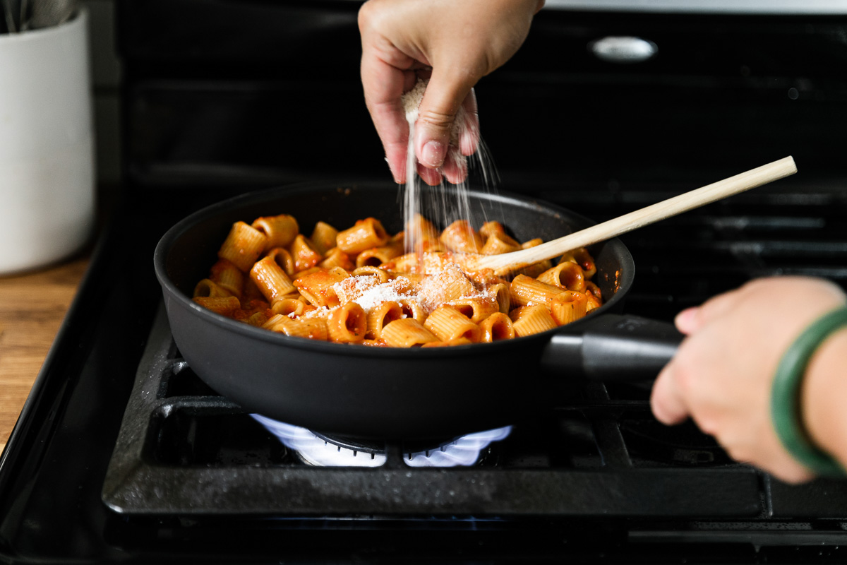 Cooked DeLallo Mezzi Rigatoni in a skillet tossed and combined with pasta sauce a woman's hands sprinkle parmesan cheese atop of the pasta to thicken up the sauce and help to create a cohesive dish. A wooden spoon rests inside the skillet and the skillet sits atop a gas stovetop range.