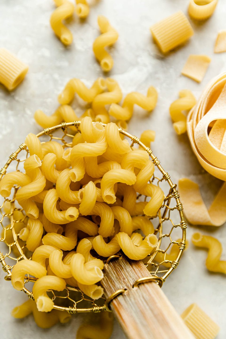 An overhead shot of a spider strainer filled with raw uncooked cavatappi pasta from DeLallo Foods. The spider strainer rests on a creamy white plaster surface. Pieces of cavatappi, Mezzi Rigatoni, and pappardelle are scattered around the spider strainer.