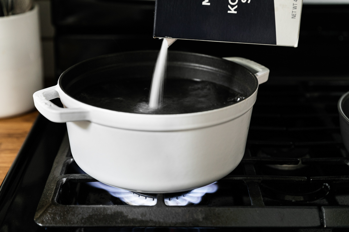 A woman's hand holds a box of Morton Coarse Kosher Salt and pours the salt into a dutch oven filled with boiling water to help cook pasta perfectly. The white dutch sits atop a gas stovetop.