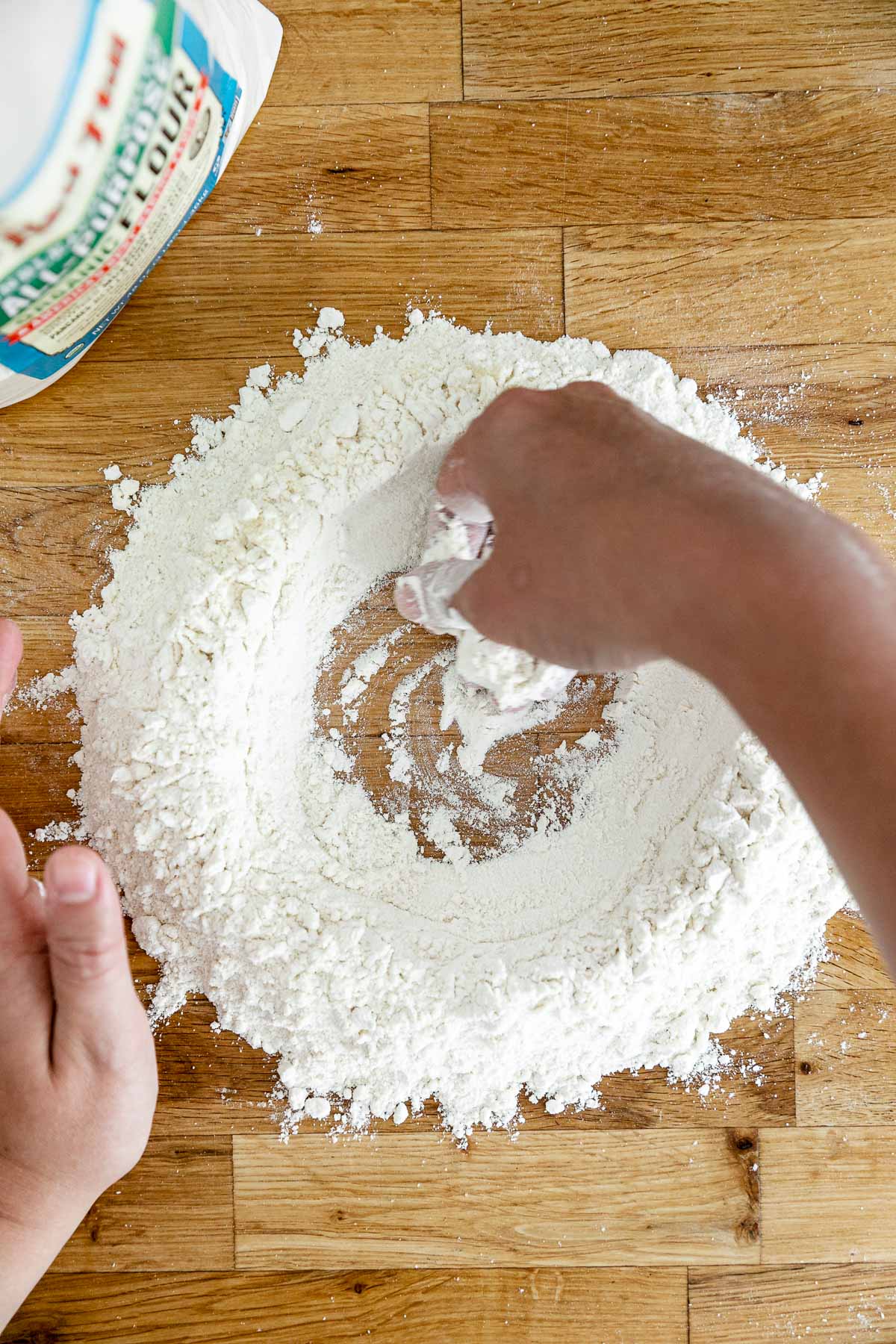 How to Make Homemade Pasta, Step 1: Mixing the flours. An overhead shot of Jess of Plays Well With Butter creating a large well in the center of a flour mixture with her hands. The well of flour sits atop a butcher block counter top and will be used to create fresh pasta dough.