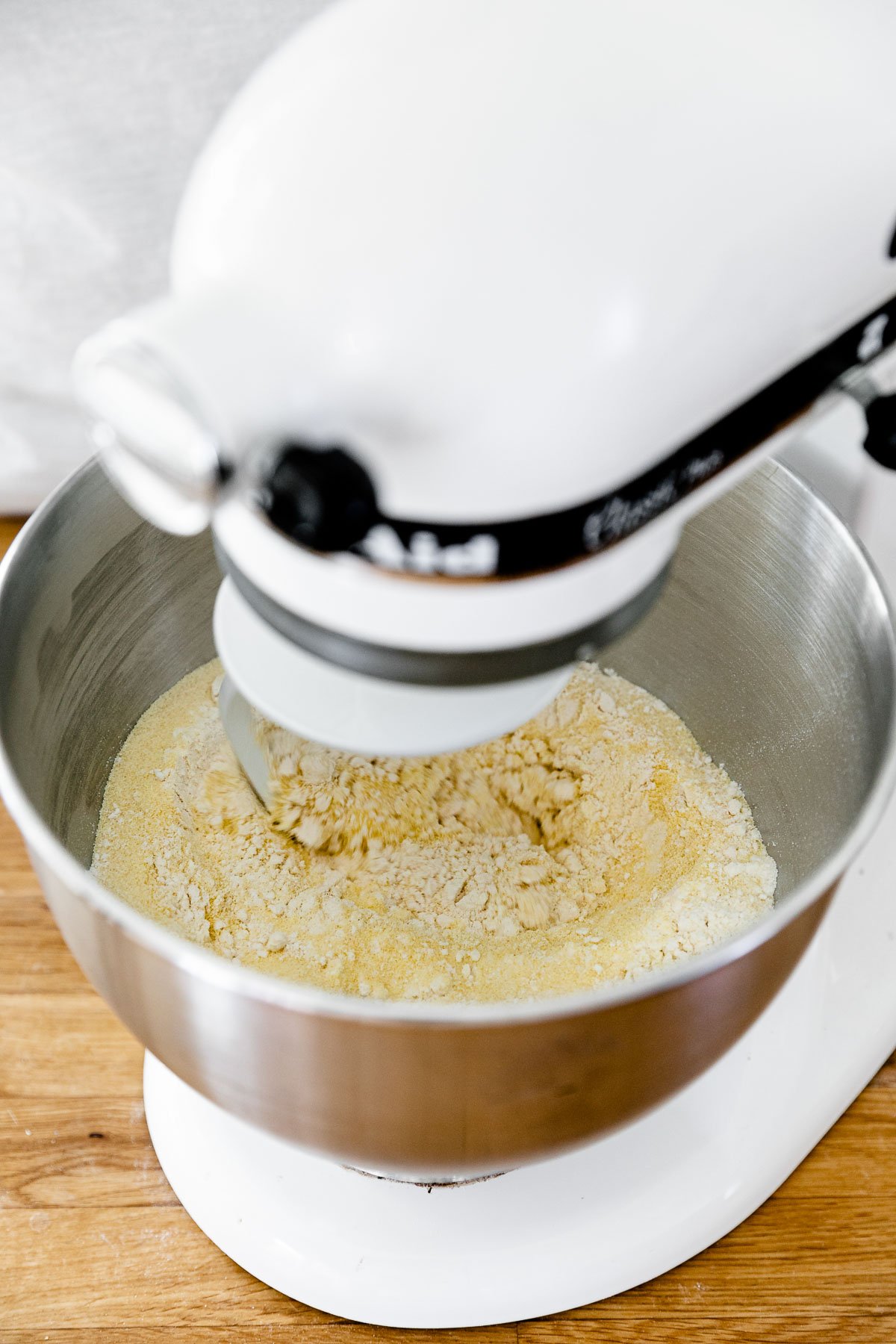How to Make Fresh Pasta Dough with a Stand Mixer, Step 2: Mix the pasta dough. A KitchenAid Stand Mixer with the paddle attachment connected is turned on and being used knead fresh pasta dough that is forming within the mixing bowl. The stand mixer rests atop a butcher block countertop.