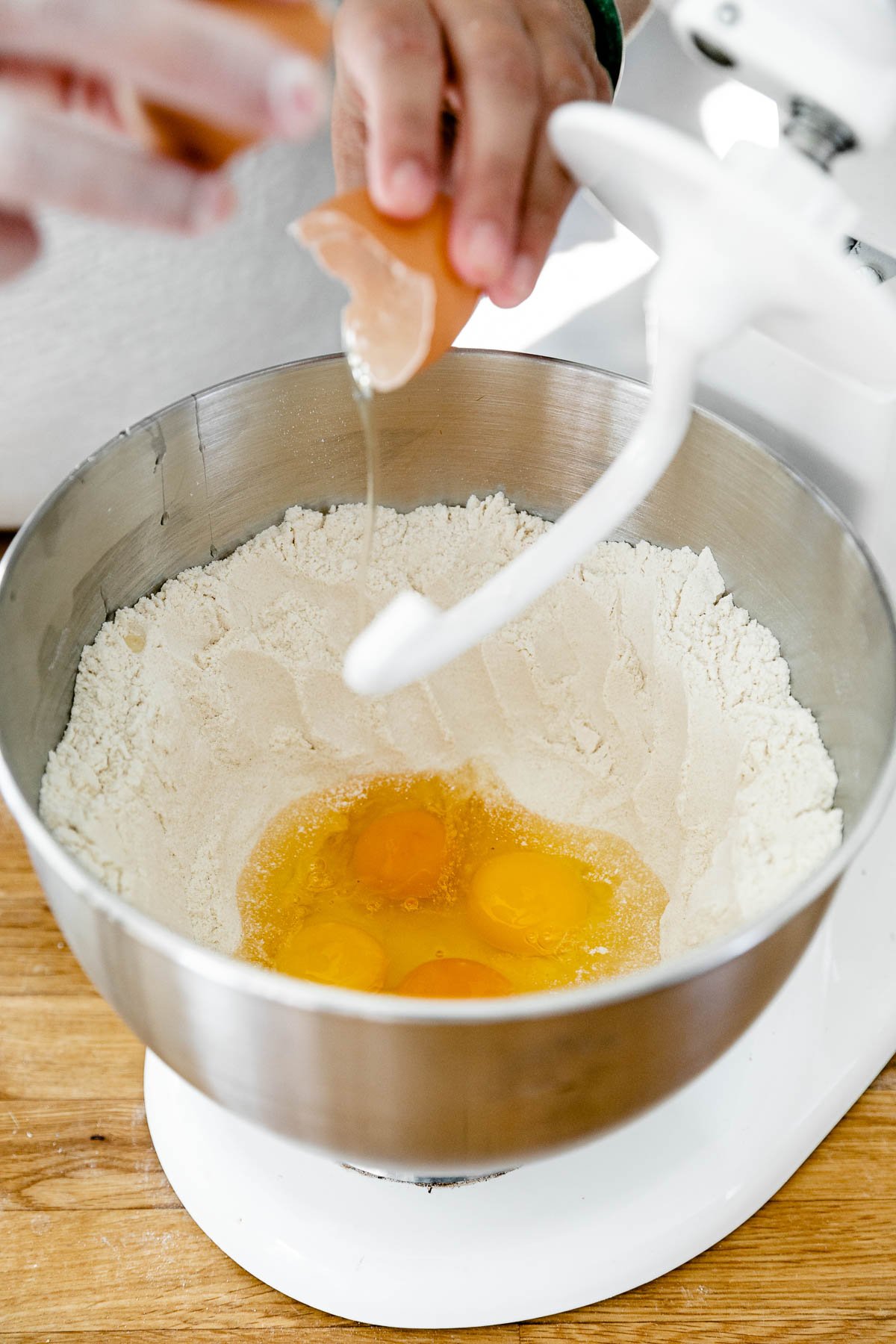 How to Make Fresh Pasta Dough with a Stand Mixer, Step 2: Mix the pasta dough. A woman's hand cracks a single egg into a well of flour formed inside mixing bowl of a white KitchenAid Stand Mixer. The dough attachment for the stand mixer is attached and is in the upright position. A few other eggs have already been added to the well of flour. The stand mixer rests atop a butcher block countertop.