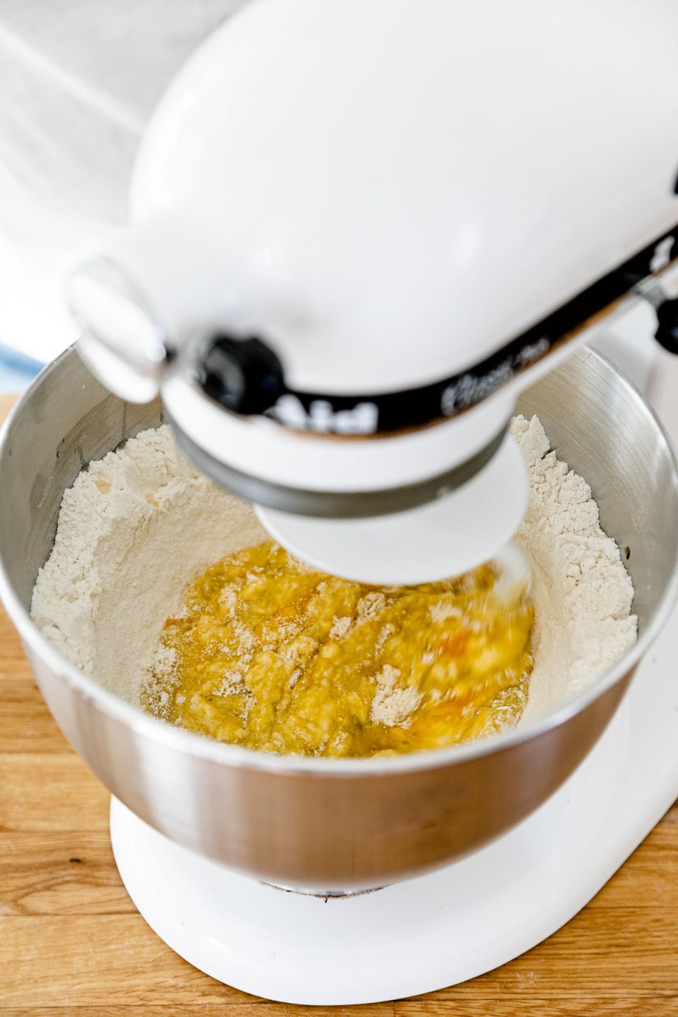 How to Make Fresh Pasta Dough with a Stand Mixer, Step 2: Mix the pasta dough. A KitchenAid Stand Mixer with the dough hook attachment connected is turned on and being used to mix wet ingredients into a well of flour to create fresh pasta dough. The stand mixer rests atop a butcher block countertop.