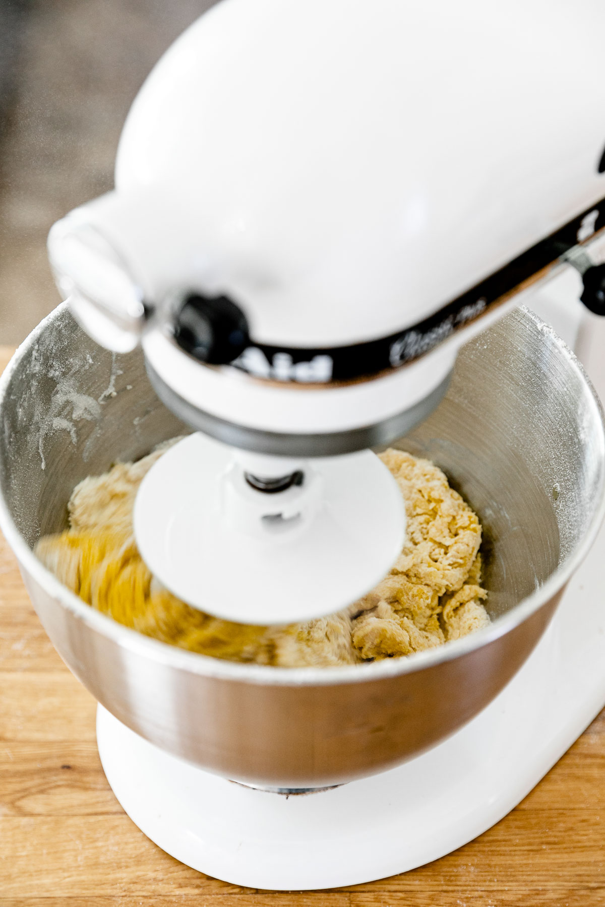 How to Make Fresh Pasta Dough with a Stand Mixer, Step 2: Mix the pasta dough. A KitchenAid Stand Mixer with the dough hook attachment connected is turned on and being used knead fresh pasta dough that is forming within the mixing bowl. The stand mixer rests atop a butcher block countertop.