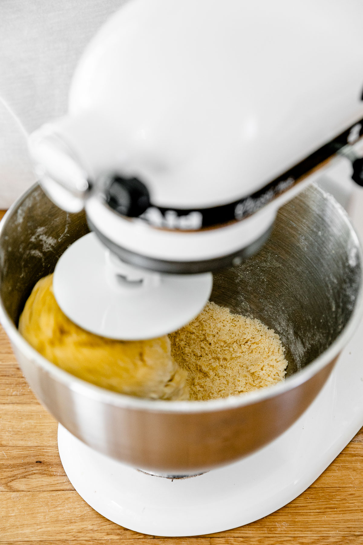 How to Make Fresh Pasta Dough with a Stand Mixer, Step 2: Mix the pasta dough. A KitchenAid Stand Mixer with the dough hook attachment connected is turned on and being used knead fresh pasta dough that is forming within the mixing bowl. The stand mixer rests atop a butcher block countertop.