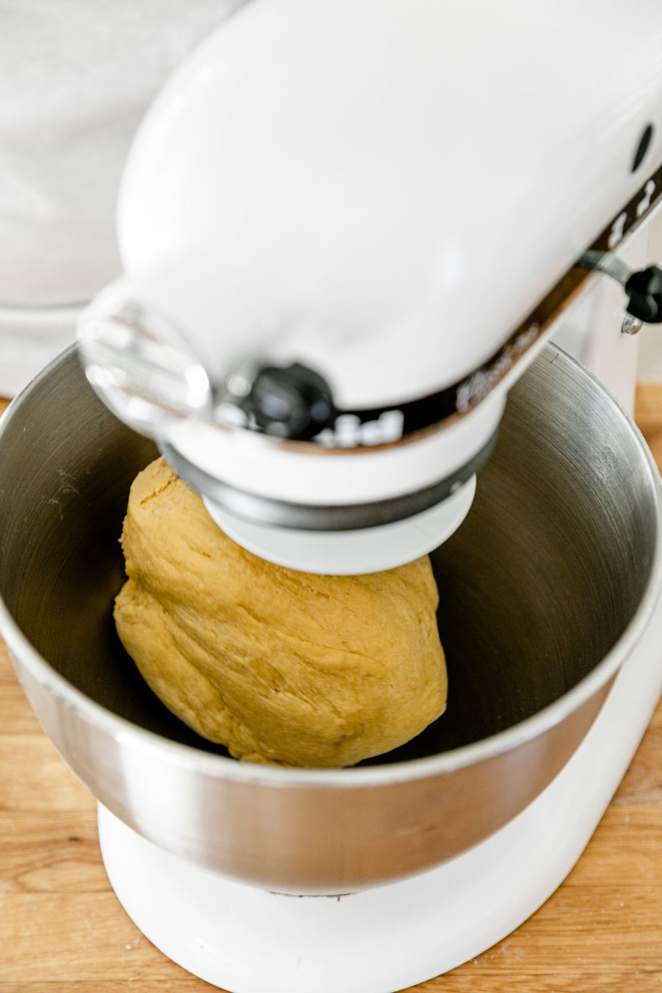How to Make Fresh Pasta Dough with a Stand Mixer, Step 2: Mix the pasta dough. A KitchenAid Stand Mixer with the dough hook attachment connected is turned on and being used to knead fresh pasta dough that is forming within the mixing bowl. The stand mixer rests atop a butcher block countertop.