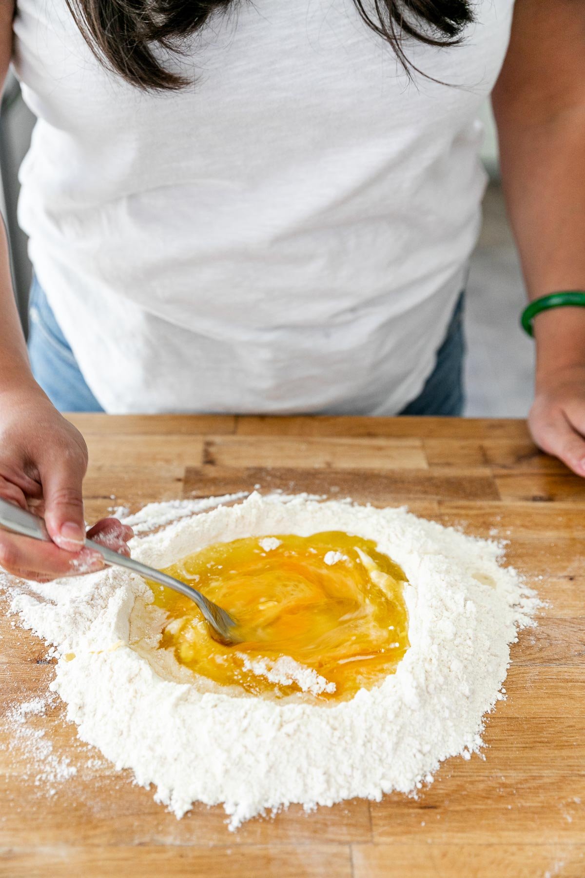 How to Make Homemade Pasta, Step 2: Mixing the Pasta Dough. Jess of Plays Well With Butter uses a fork to break up the eggs & whisk the wet ingredients inside of the well created with flour to make fresh pasta dough. The ingredients sit atop a clean butcher block countertop.