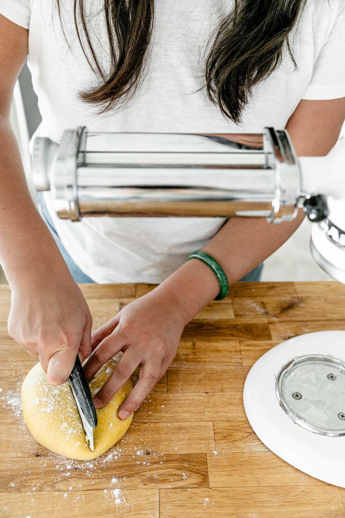 Jess of Plays Well With Butter uses a bench scraper to divide rested fresh pasta dough into pieces prior to rolling it out using a pasta attachment on her KitchenAid Stand Mixer. The pasta dough has been lightly dusted with flour and sits atop a butcher block countertop.