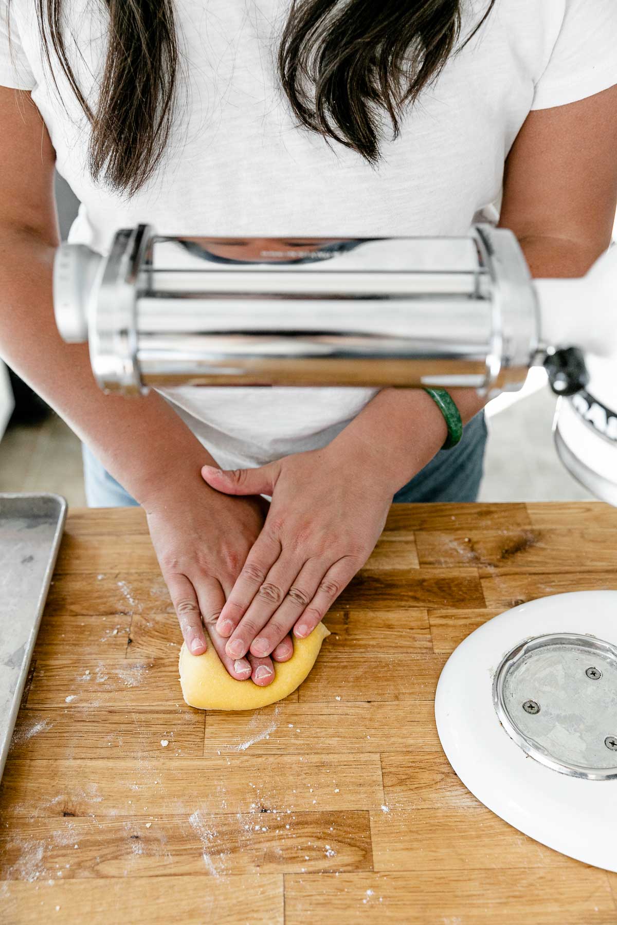 Jess of Plays Well With Butter uses her hands to press homemade pasta dough into a thin and even layer. The dough sits atop a butcher block countertop that has been lightly dusted with flour. A KitchenAid Stand Mixer with a pasta rolling attachment is set up to be used to roll out the pasta dough and an aluminum baking sheet also sits on top of the countertop.