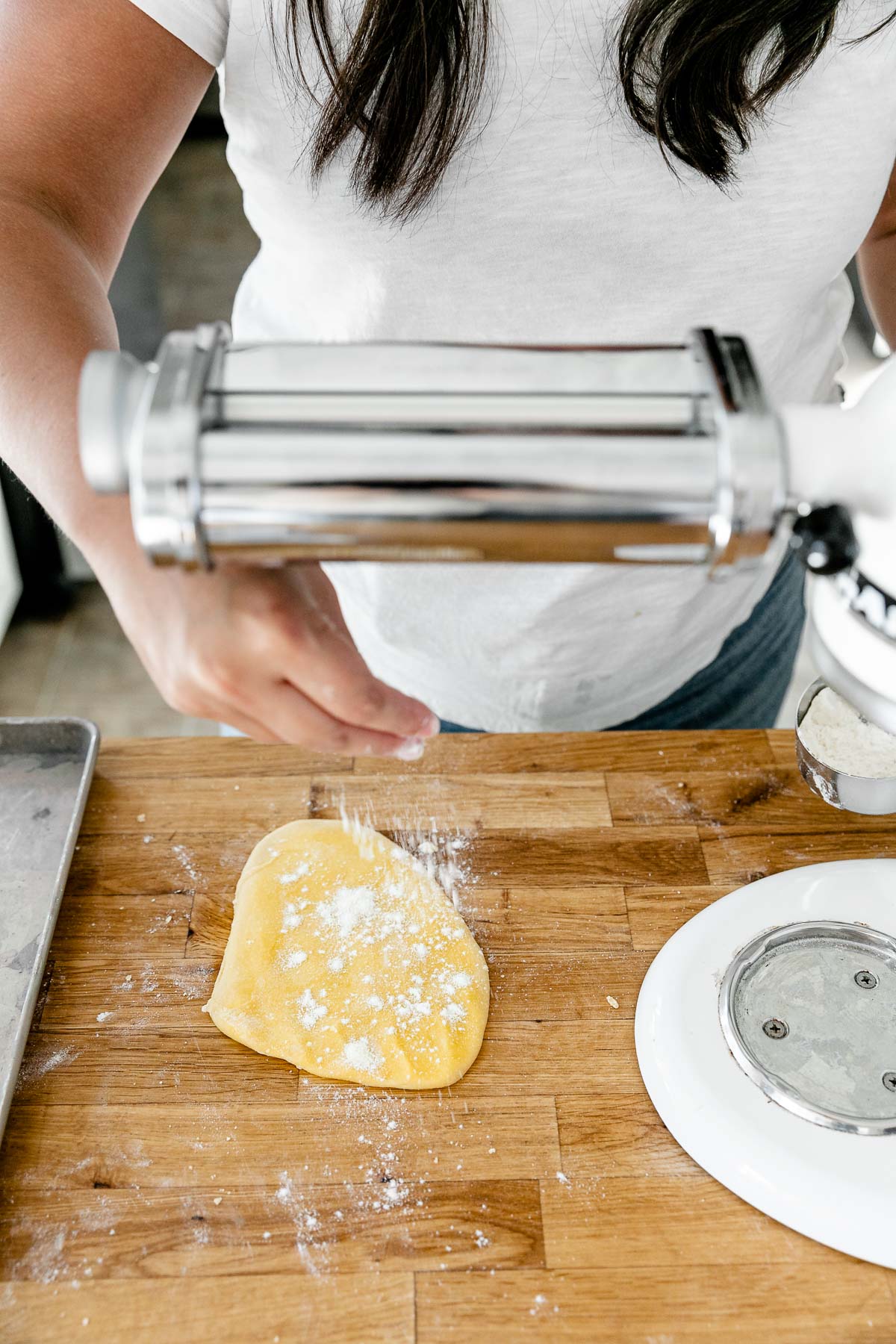 Jess of Plays Well With Butter uses her hands to lightly dust a piece of homemade pasta dough that has been pressed into a thin and even layer and rests atop a butcher block countertop. ​A KitchenAid Stand Mixer with a pasta rolling attachment is set up to be used to roll out the pasta dough and an aluminum baking sheet also sits on top of the countertop.