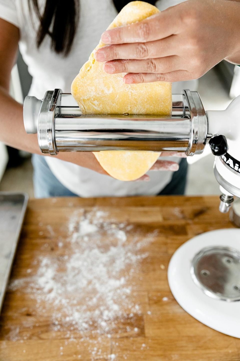 Jess of Plays Well With Butter uses her hands to feed a piece of homemade pasta dough through a pasta roller attachment connected to a KitchenAid Stand Mixer. The Stand Mixer rests atop a butcher block countertop. The countertop has been lightly dusted with flour and an aluminum baking sheet also sits on top of the countertop.