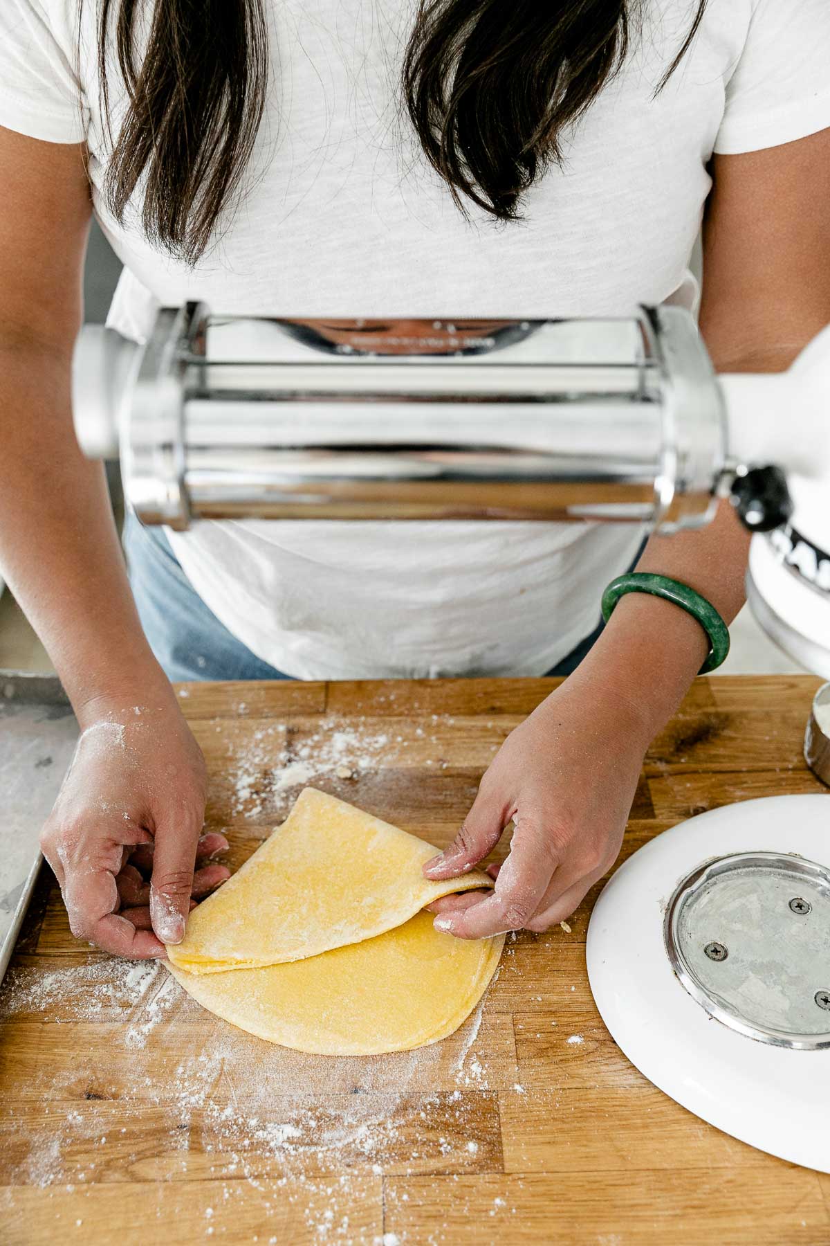 Jess of Plays Well With Butter uses her hands to fold homemade pasta dough in half atop a flour dusted butcher block countertop. ​A KitchenAid Stand Mixer with a pasta rolling attachment is set up to be used to roll out the pasta dough and an aluminum baking sheet also sits on top of the countertop.