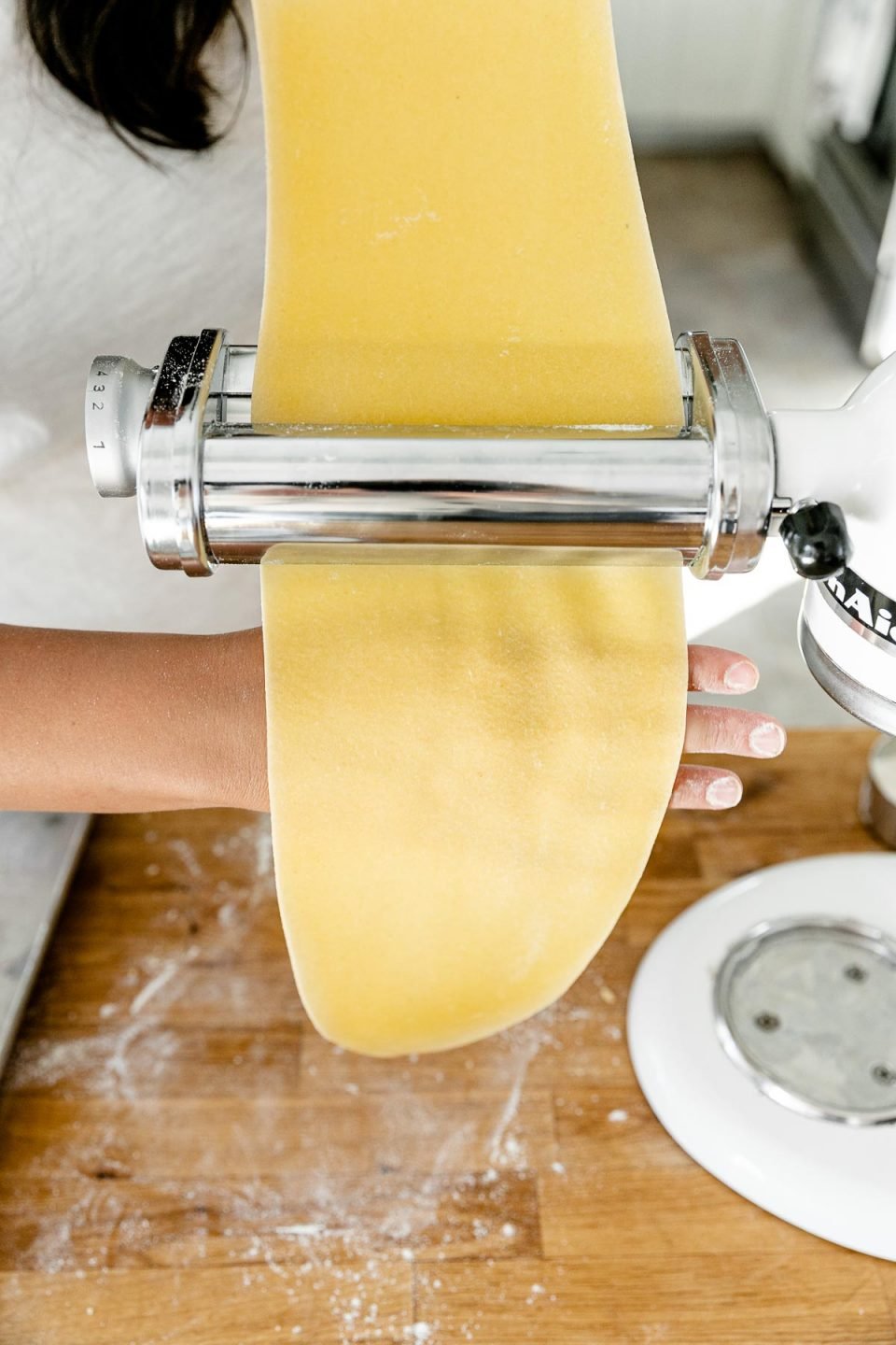 Jess of Plays Well With Butter uses her hands to feed a piece of homemade pasta dough through a pasta roller attachment connected to a KitchenAid Stand Mixer. Her top hand holds the sheet of dough that is being rolled out while the back of her bottom hand is used to catch the pasta dough. The Stand Mixer rests atop a butcher block countertop. The countertop has been lightly dusted with flour and an aluminum baking sheet also sits on top of the countertop.