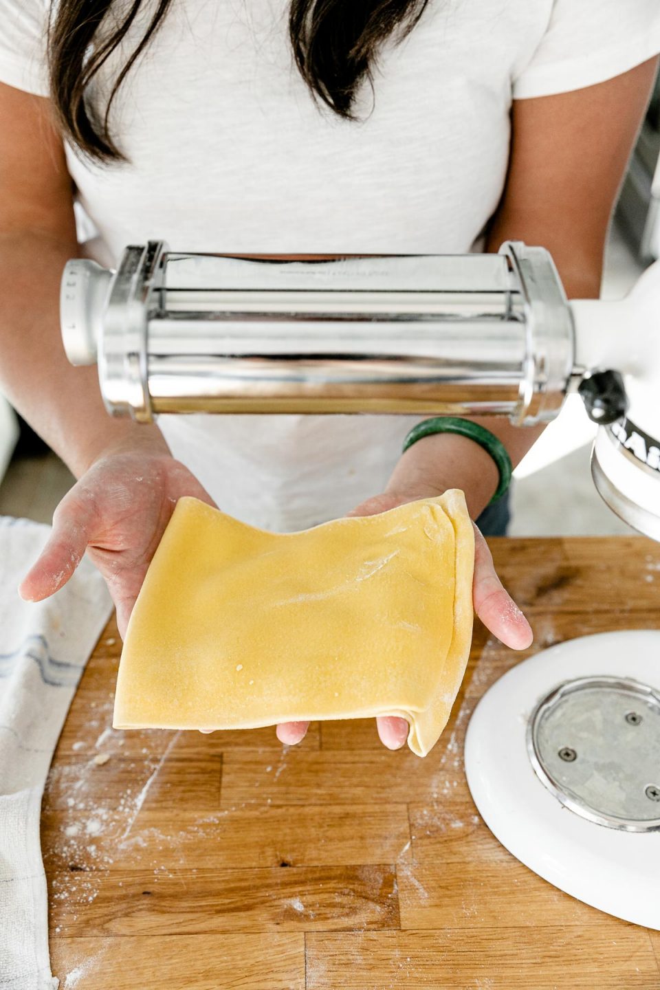 Jess of Plays Well With Butter holds a whole sheet of rolled pasta dough with both hands over a butcher block countertop. ​A KitchenAid Stand Mixer with a pasta rolling attachment is set up to be used to roll out the pasta dough and an aluminum baking sheet also sits on top of the countertop.
