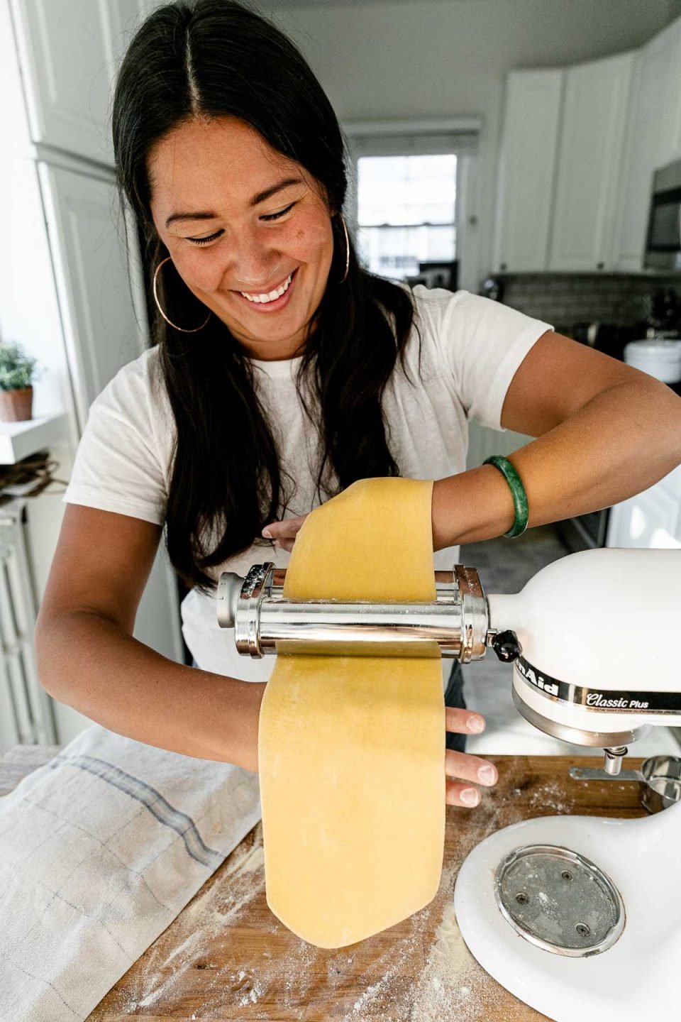 Jess of Plays Well With Butter smiles and looks downward as she rolls out pasta dough using a KitchenAid pasta rolling attachment. She uses her hands to feed a piece of homemade pasta dough through a pasta roller attachment connected to a KitchenAid Stand Mixer. Her top hand holds the sheet of dough that is being fed through the attachment while the back of her bottom hand is used to catch the pasta dough sheet that is being formed. The Stand Mixer rests atop a butcher block countertop. The countertop has been lightly dusted with flour and an aluminum baking sheet also sits on top of the countertop and is covered by a kitchen towel.