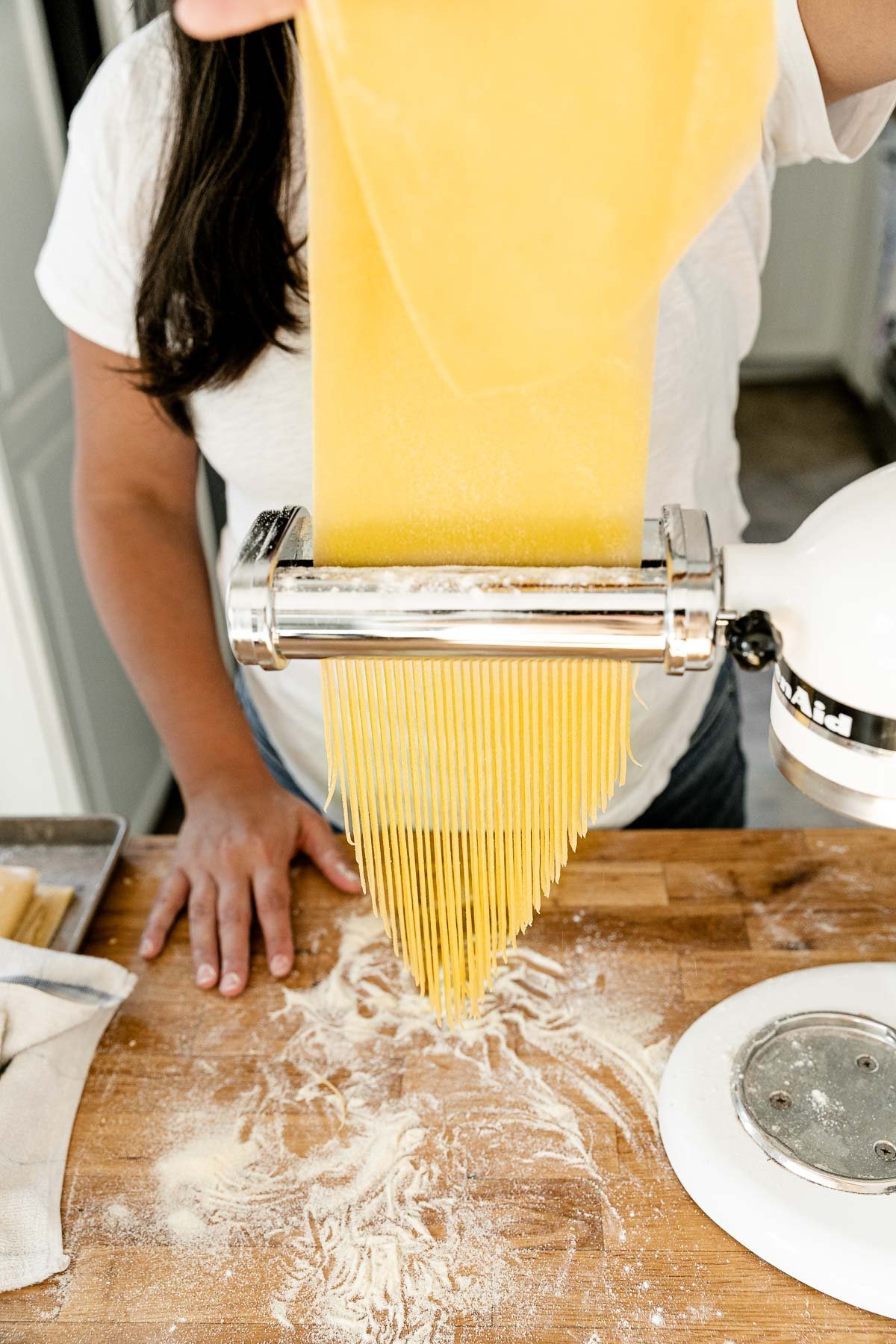 Jess of Plays Well With Butter feeds a large sheet of fresh pasta dough through a KitchenAid pasta cutting attachment connected to a Stand Mixer. As the pasta dough is fed through the cutter, the dough is cut into single strands of spaghetti. The Stand Mixer rests atop a butcher block countertop. The countertop has been lightly dusted with flour and an aluminum baking sheet also sits on top of the countertop and is covered by a kitchen towel.