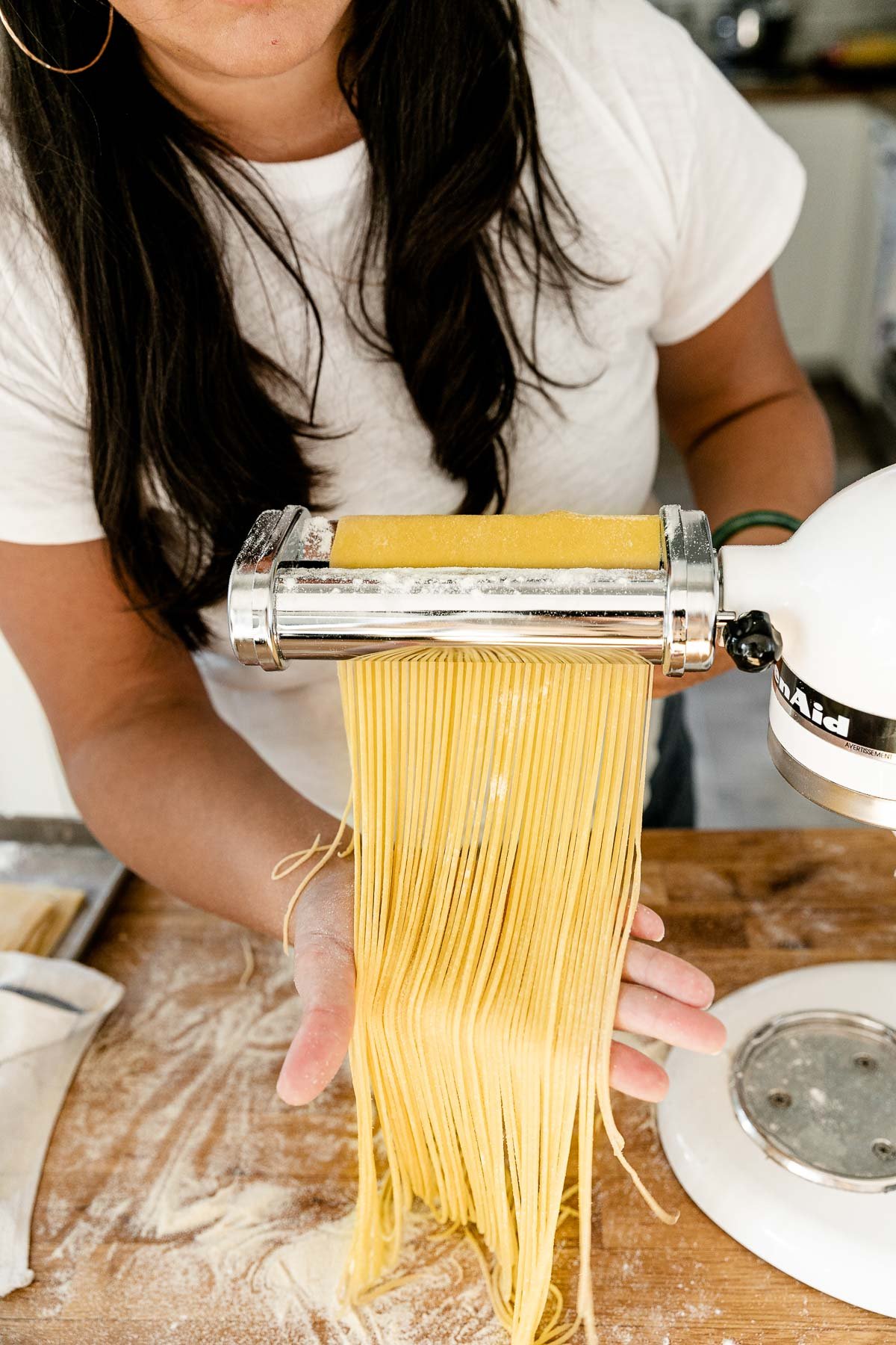 Jess of Plays Well With Butter feeds a large sheet of fresh pasta dough through a KitchenAid pasta cutting attachment connected to a Stand Mixer. As the pasta dough is fed through the cutter, the dough is cut into single strands of spaghetti. Her right hand catches the spaghetti noodles and lifts them up slightly with a flat palm to display the cut noodles and allow them to rest gently on the countertop. The Stand Mixer rests atop a butcher block countertop. The countertop has been lightly dusted with flour and an aluminum baking sheet also sits on top of the countertop and is covered by a kitchen towel.