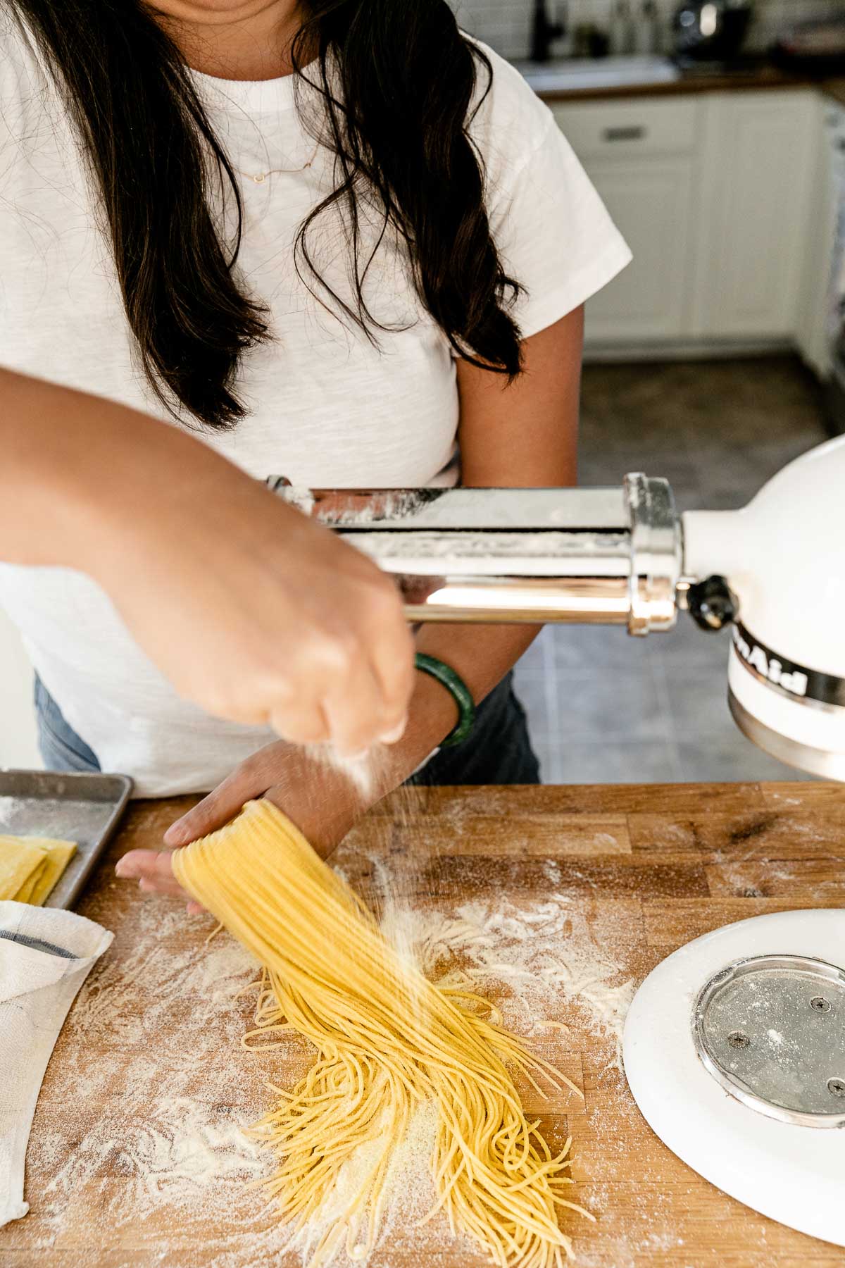 A woman loosely holds strands of fresh pasta cut into spaghetti in one hand as the bottom of the strands rest on a butcher block countertop lightly dusted with flour. ​A KitchenAid Stand Mixer with a pasta rolling attachment is set up to be used to roll out the pasta dough and an aluminum baking sheet also sits on top of the countertop.