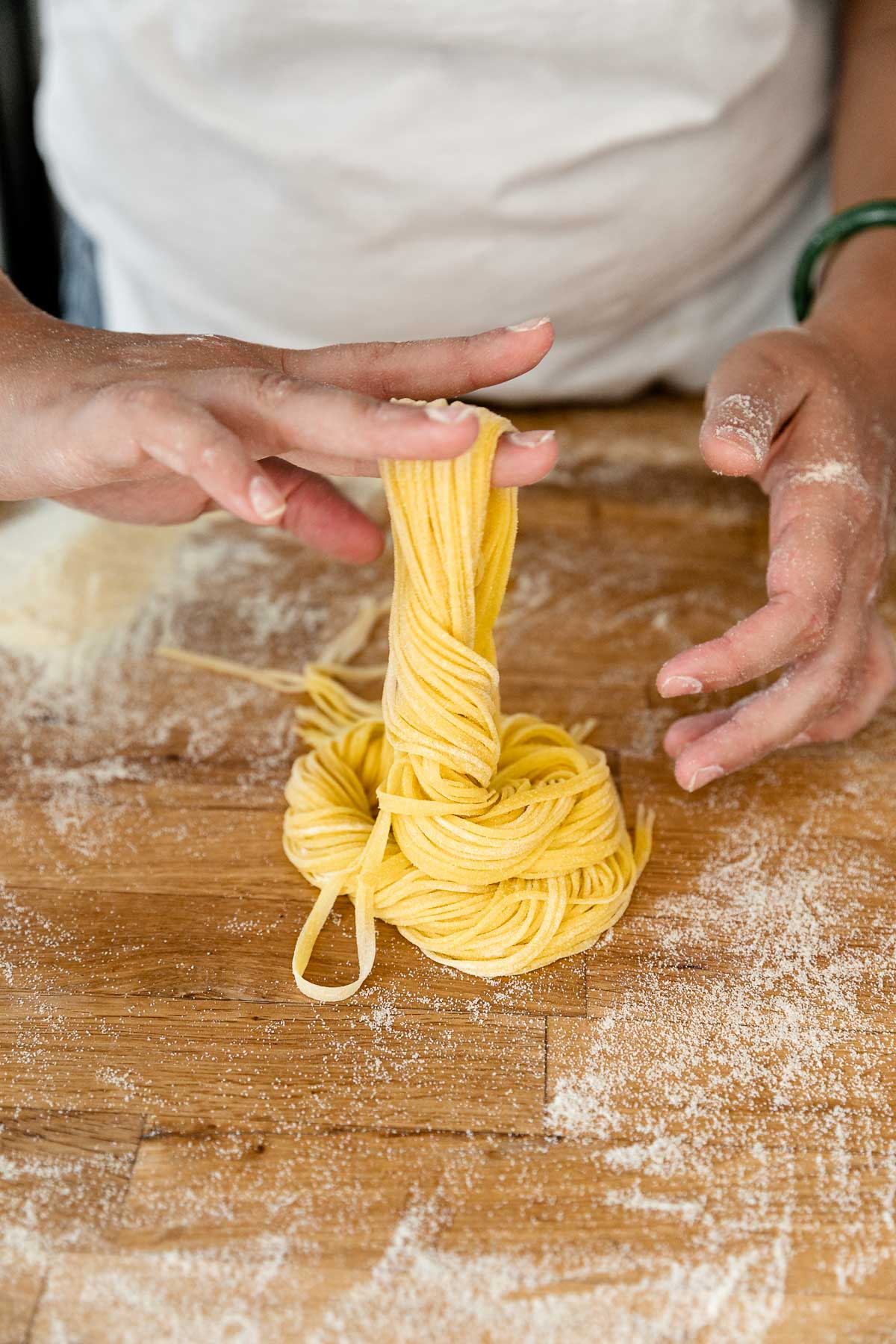 A side angle shot of a woman's hands forming a bundle of fresh pasta from homemade pasta dough cut into spaghetti noddles. The noodles are draped across her middle finger as she twists and lets the noodles fall into a loose bundle. The bundle rests atop a butcher block countertop that is dusted with semolina flour.